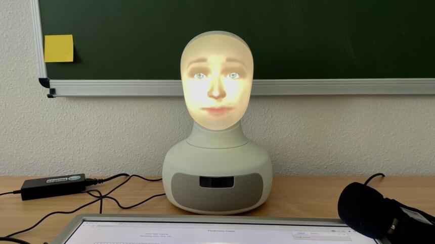 Eine Büste eines Roboters mit einem menschenähnlichen Gesicht vor einer Schultafel. Vor ihm liegt ein Mikrofon.